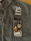 Close-up of a dark charcoal gray denim jacket showing a pocket with a 'Broken Promises' patch and two enamel pins, alongside white embroidered script reading 'The last time' on the chest.