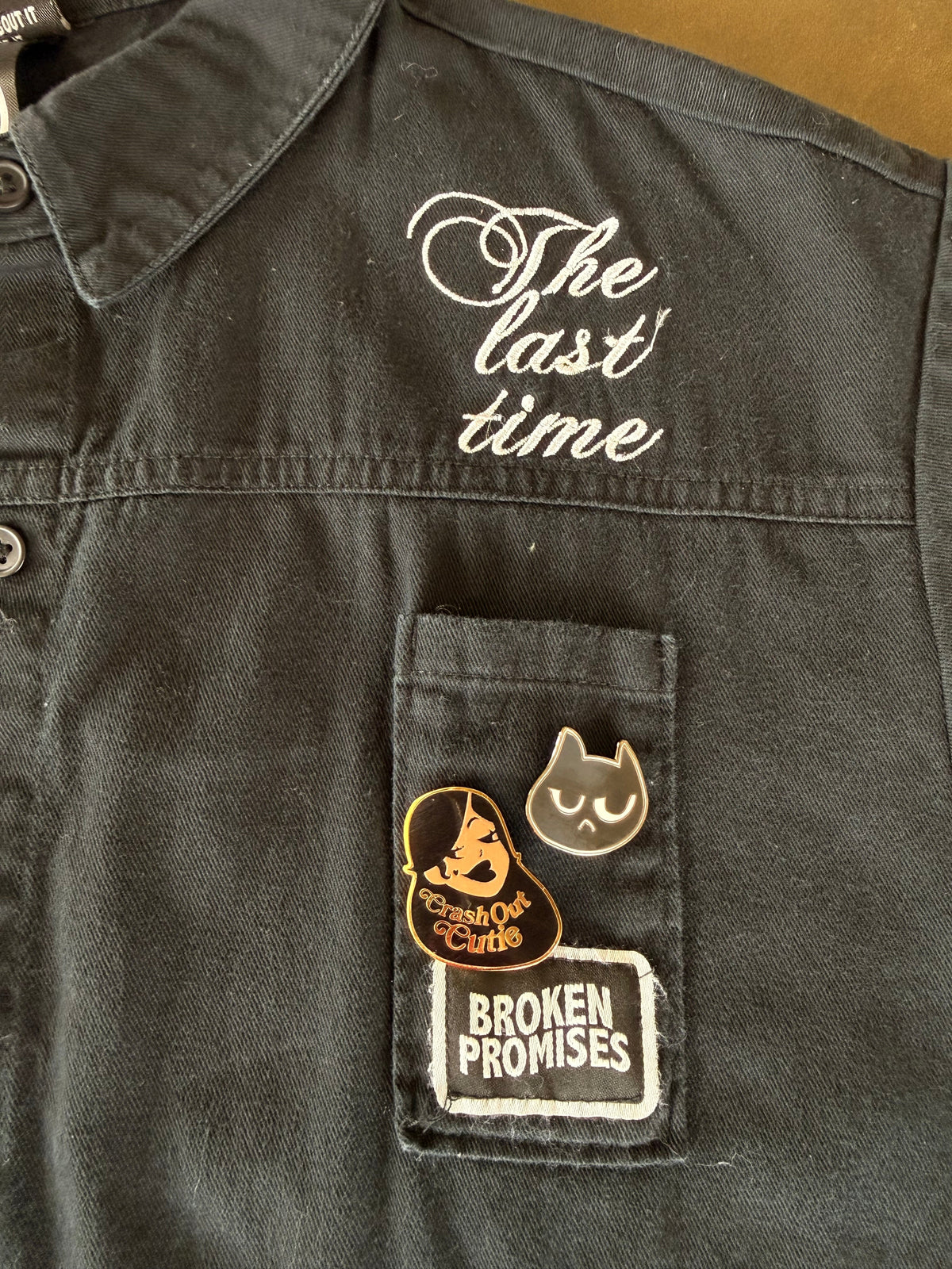 Close-up of a dark charcoal gray denim jacket showing a pocket with a 'Broken Promises' patch and two enamel pins, alongside white embroidered script reading 'The last time' on the chest.