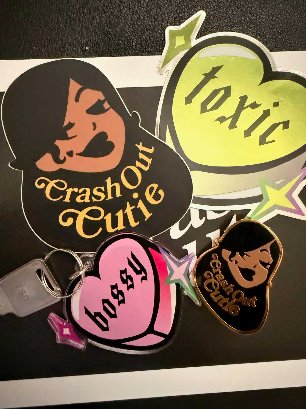 A set of die-cut acrylic keychain charms and stickers featuring cartoon character graphics and text including 'Crash Out Cutie', 'toxic', and 'bossy'.