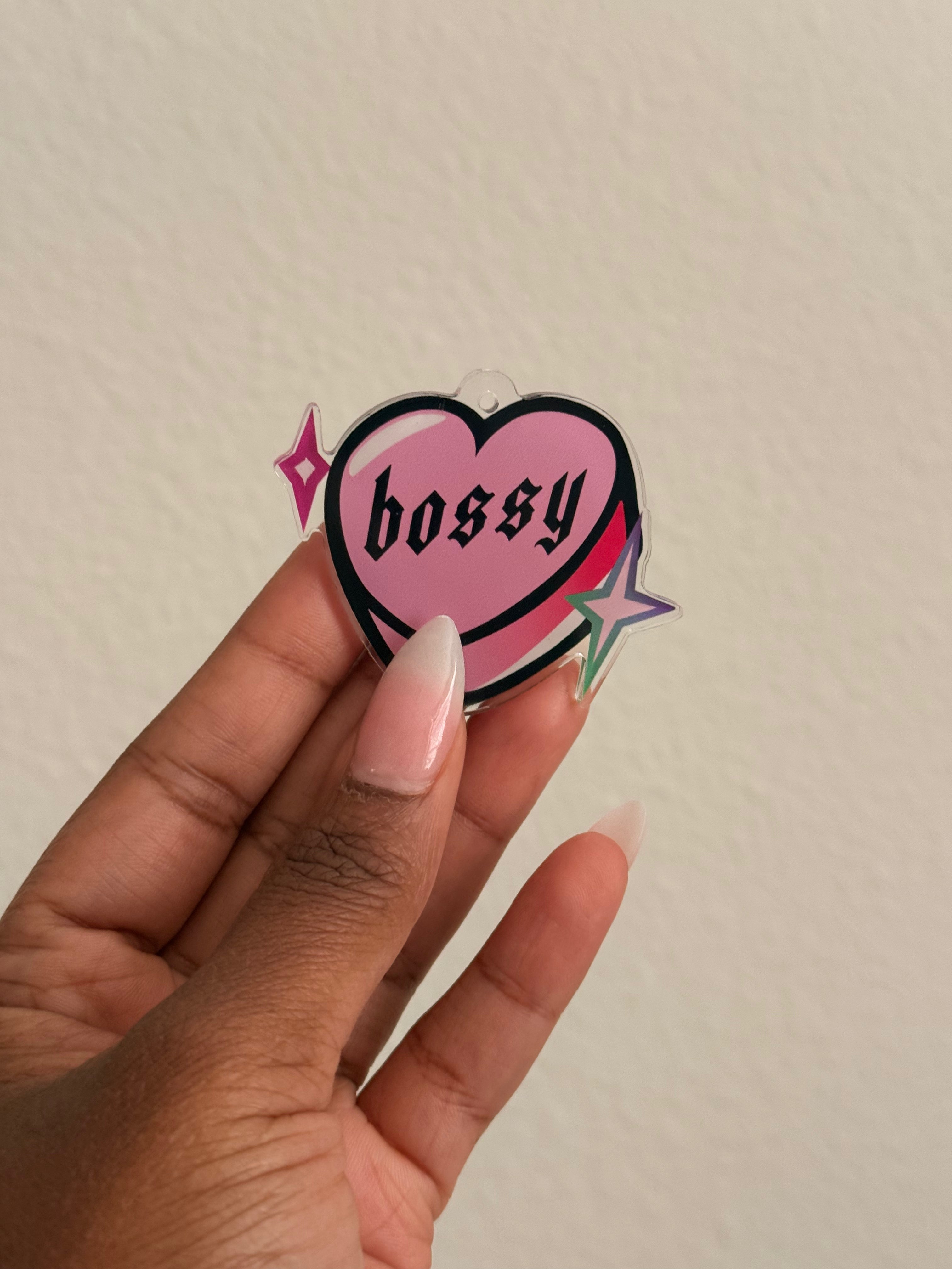 Heart-shaped sticker with 'bossy' text held by a hand against a plain background